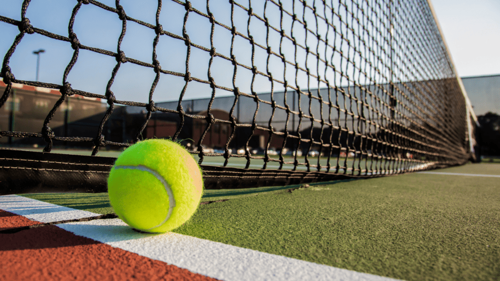 Close up of a tennis ball on tennis turf with a net and court lines in the background 1