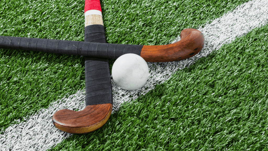 Close up of two field hockey sticks crossed over a white line on field hockey turf with a white hockey ball placed between them