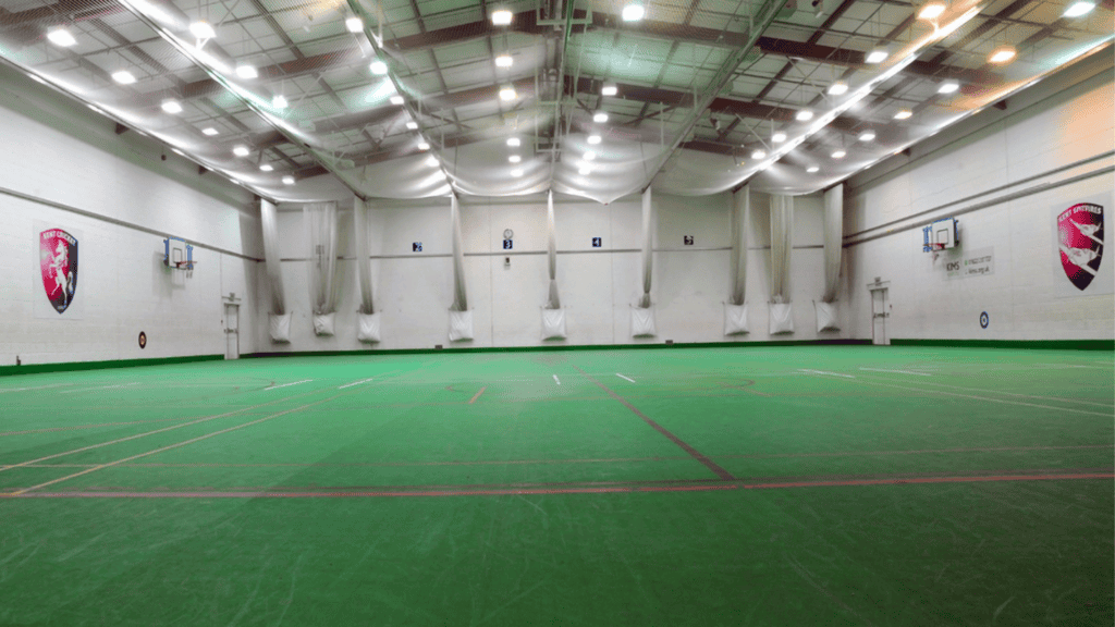 Indoor sports facility with green turf surrounded by cricket nets and basketball hoops