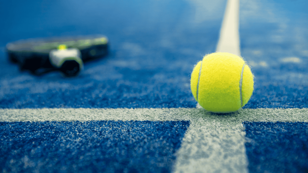 Yellow tennis ball on a blue tennis court with a blurred racket in the background showing the courts baseline