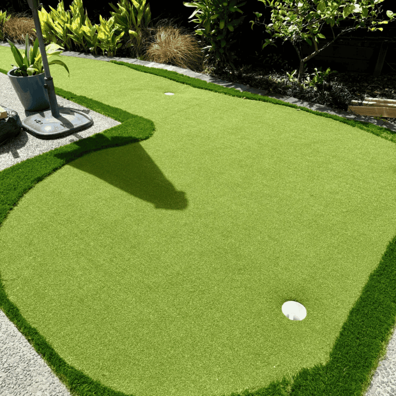 Outdoor 1 small backyard golf putting green with synthetic grass surrounded by plants and umbrella for home golf practice