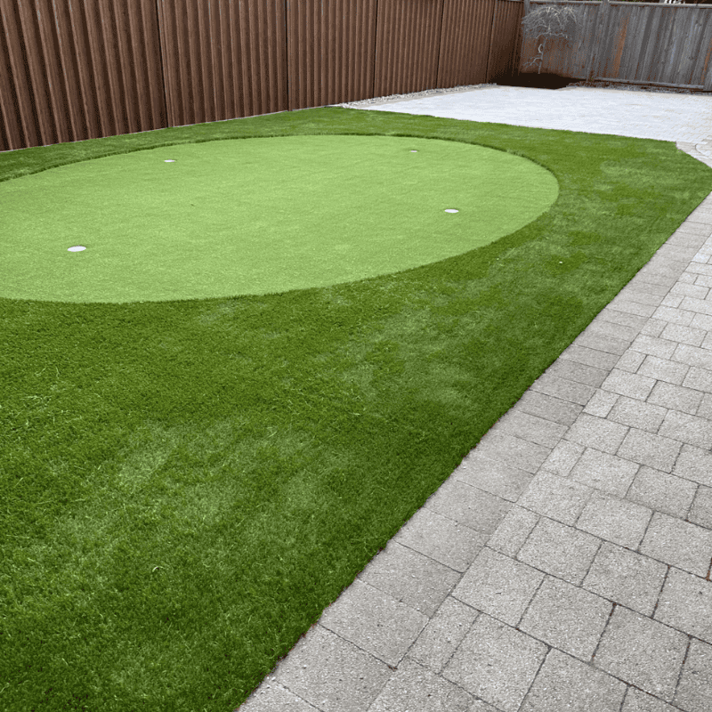small backyard putting green with synthetic grass surrounded by fence and paver walkway for home golf practice