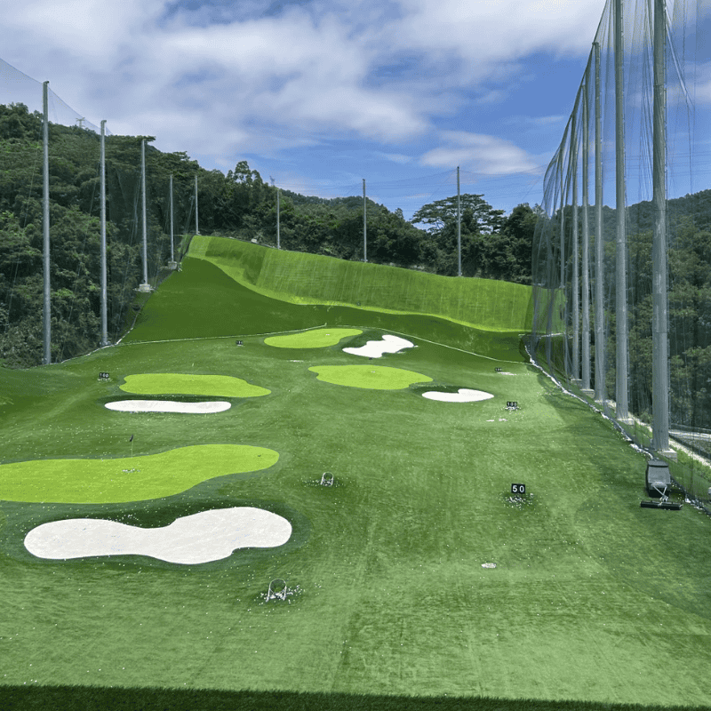 Outdoor 2 synthetic grass driving range with multiple holes surrounded by tall netting and bright sky above