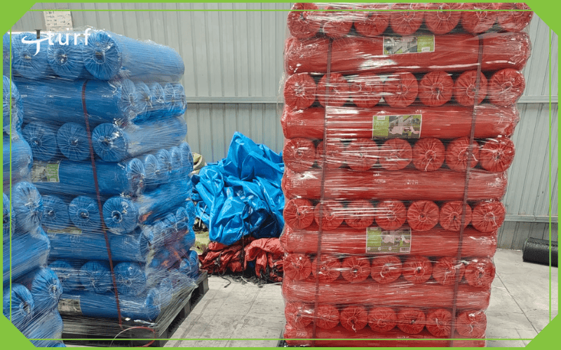 Rollos paletizados de césped artificial en envoltorio azul y rojo listos para envío