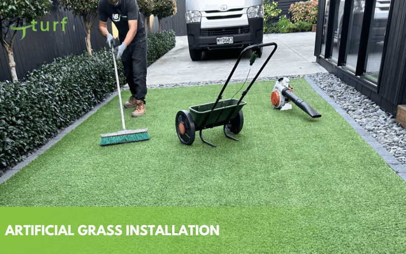 Worker installing artificial grass using broom and spreader tools in driveway area