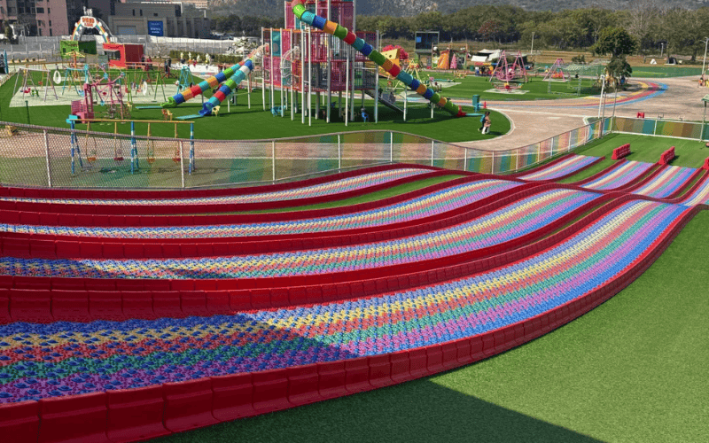 Acolchado de césped para áreas de juego 101 para superficies más seguras y suaves 1 Largos toboganes de césped arcoíris en un parque infantil al aire libre con césped artificial