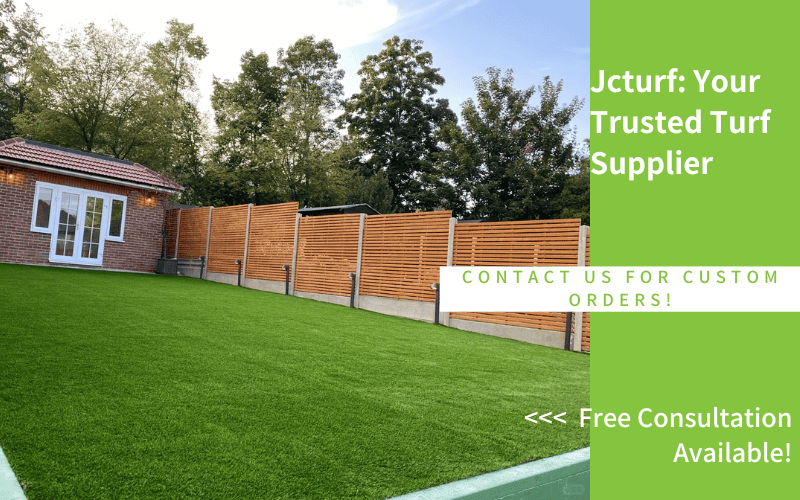 A green artificial turf lawn is shown next to a brick building, surrounded by wooden fencing and trees in the background.