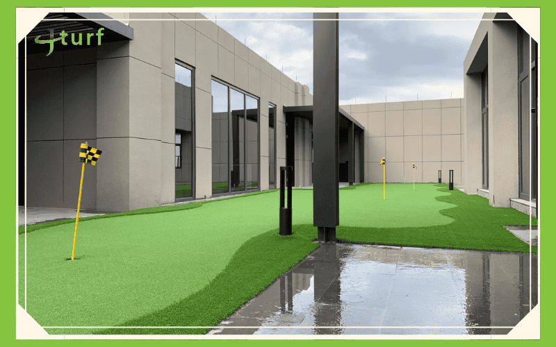 A modern outdoor space featuring artificial turf with putting greens and yellow flags, framed by large windows and gray buildings.