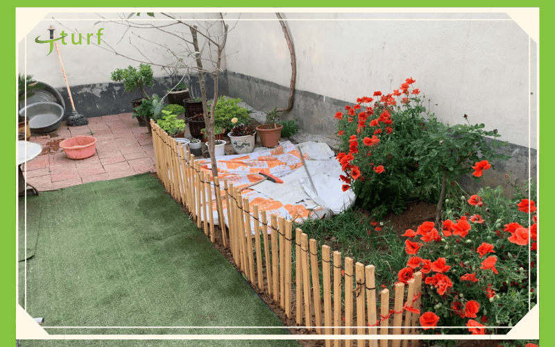 Un área de jardín con flores de color naranja vibrante, plantas en macetas y una cerca de bambú, con un trozo de césped verde.