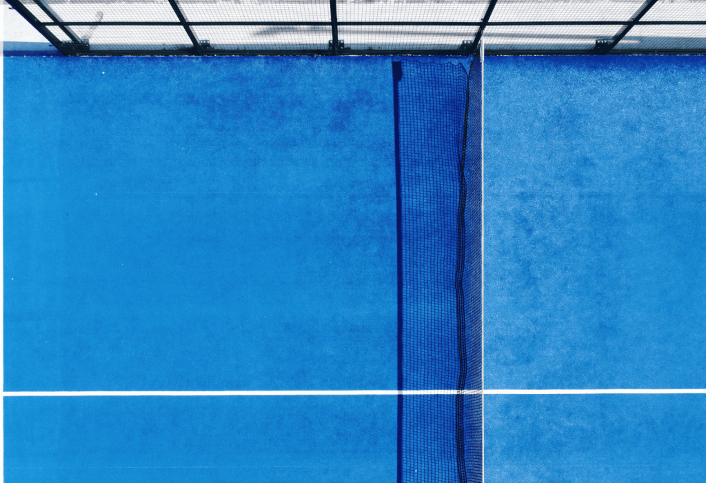 Aerial view of a padel court with blue turf, featuring a white line marking the center and a black net along the side Aerial view of a padel court with blue turf, featuring a white line marking the center and a black net along the side