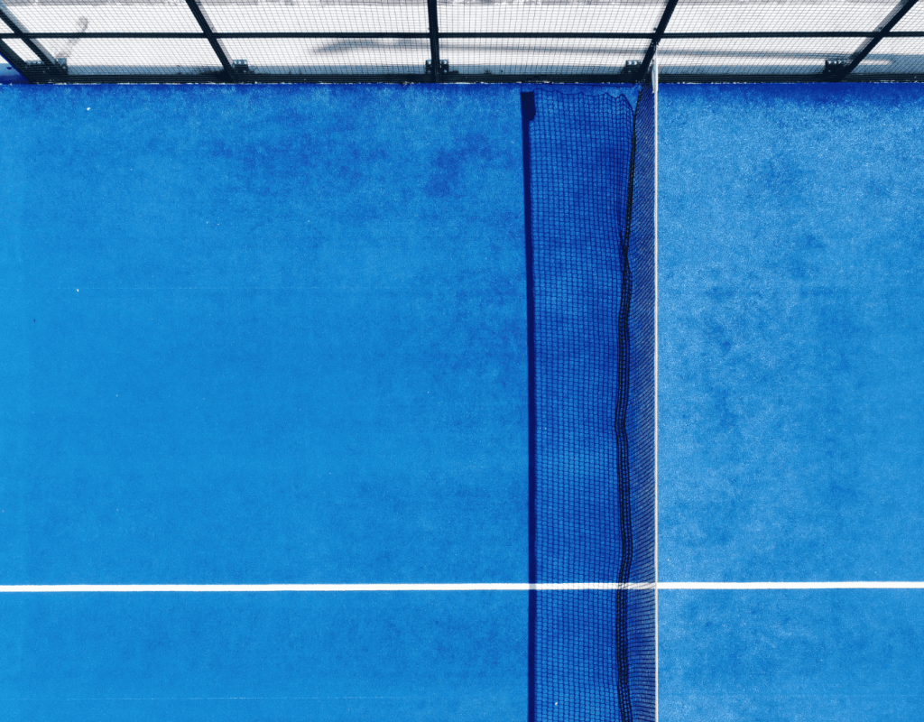Aerial view of a padel court with blue turf, featuring a white line marking the center and a black net along the side Aerial view of a padel court with blue turf, featuring a white line marking the center and a black net along the side
