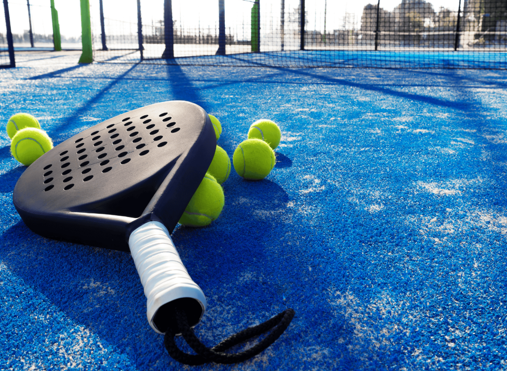 Close-up of a padel racket and several tennis balls on blue padel turf court surface with shadows from surrounding netting Close-up of a padel racket and several tennis balls on blue padel turf court surface with shadows from surrounding netting