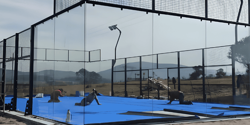 Obreros instalando una pista de pádel profesional exterior con suelo azul, rodeada de paredes de cristal y malla metálica en un entorno rural.